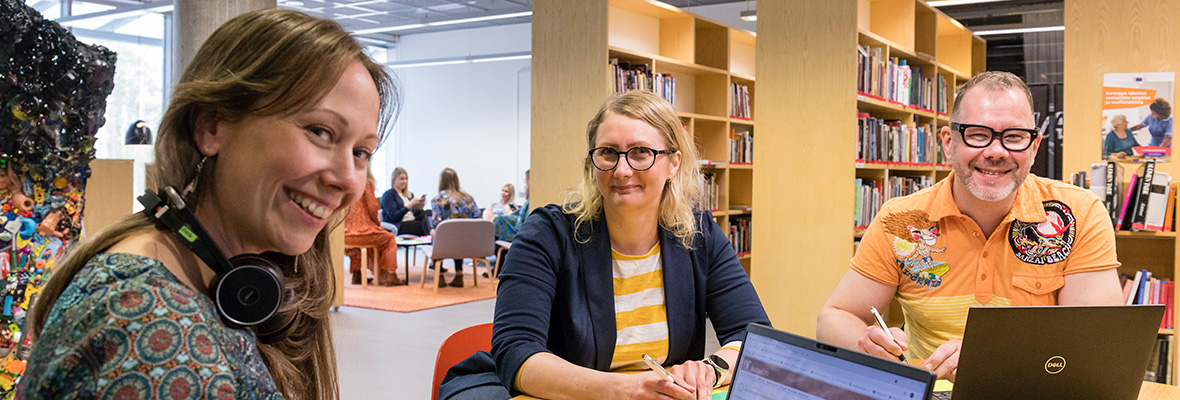 Kuvassa kaksi naista ja mies työskentelemässä Lapin yliopiston taiteiden tiedekunnan F-Hub-yhteistyöskentelytilassa, taustallaan tilataidetta ja kirjahyllyjä. Töihin yliopistoomme -verkkosivulle
