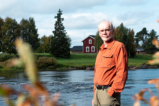 Timo Jokela seisoo Ounasjoen rannalla, takanaan virtaava joki ja puutalo. Kuva: Aki Lintumäki.