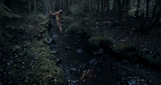 Kuva Kristiina Koskisen lyhytelokuvasta Metsän määritelmä. Kuvassa kivinen metsäpuro, aluskasvillisuutta, puita ja ihminen.