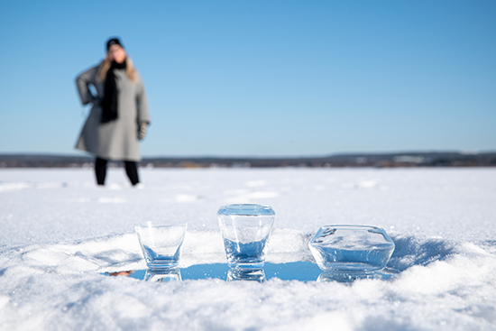 Kolme lasia aseteltuna riviin Ounasjoen jäällä, taustalla Milla Johansson seisoo matalassa lumihangessa. Kuva: Elli Alasaari.