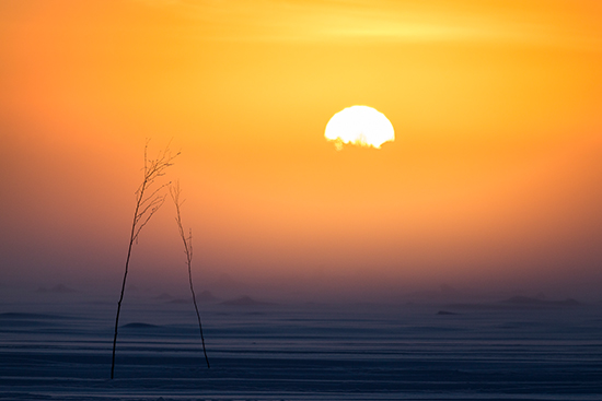 Aurinko hehkuu pakkasudun takana, etualalla lunta.