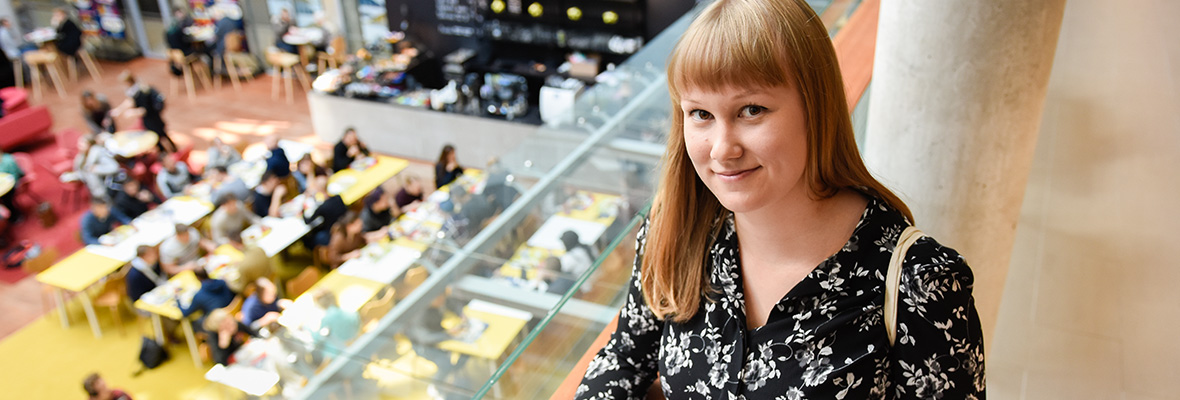 Kuvassa nainen ja hänen taustallaan Lapin yliopiston taiteiden tiedekunnan rakennuksen Agora-keskusaukio ja ihmisvilinää ravintola Petronella. Tule kylään -verkkosivuille.