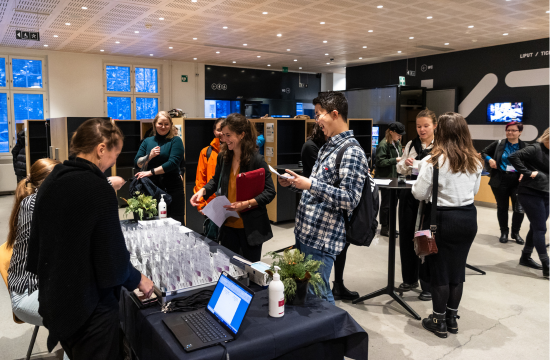 Ihmisiä rekisteröitymässä konferenssiin modernissa sisätilassa. Pöydällä on nimikortteja ja kannettava tietokone. Osallistujat näyttävät iloisilta ja keskustelevat keskenään. Taustalla näkyy pukukaappeja ja iso näyttö.