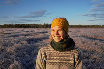 Kuvassa Sanna Hast hymyilee auringonlaskun valaisemana, taustalla jäinen suo. 