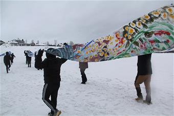 Ihmiset kulkevat lumisessa maisemassa jäällä ja nostavat käsillää npäiden yläpuolelle värikästä lakanaa.