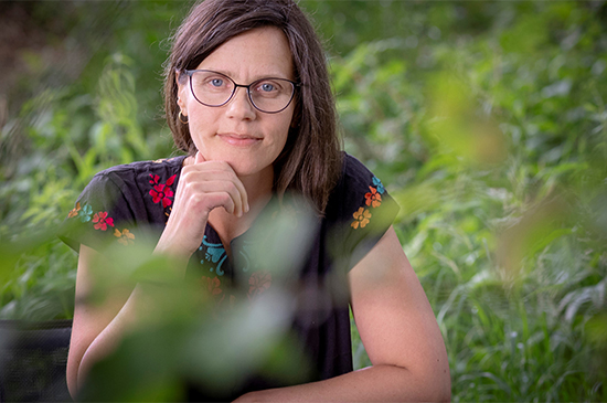 Kristiina Koskinen katsoo suoraan kohti ja nojaa leukaansa käteensä. Taustalla ja etualalla on vehreää kasvillisuutta. Kuva: Harri Pälviranta.