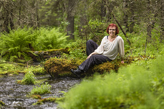 Kuvassa Eeva Rossi istuu metsässä puron varrella. 