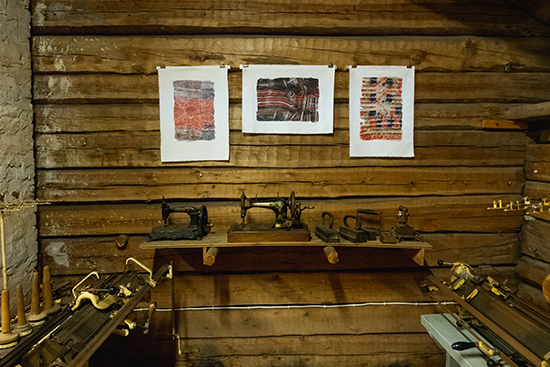 Kolme teosta ripustetuna hirsiseinälle, alapuolellaan vanhoja tarve-esineitä. Kuva: Inka Holck.