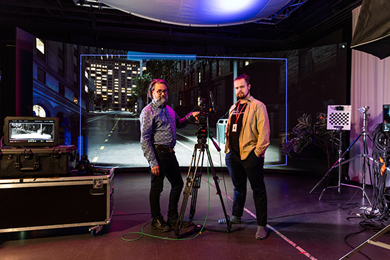 Tomi Knuutila ja Samuli Valkama seisovat virtuaalituotantostudiossa kaarevan lediseinän edessä ja molemmin puolin elokuvakameraa. Kuva: Ville Rinne.