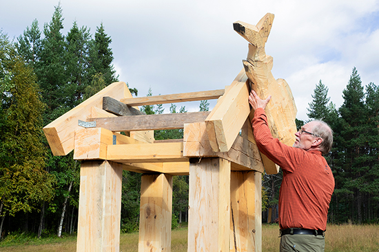 Timo Jokela pitää ylhäällä käsissään isoa kalanmuotoista puukappaletta ja asettelee sitä pientä katonharjaa muistuttavan hirsirakennelman pinnalle. Kuva: Aki Lintumäki.