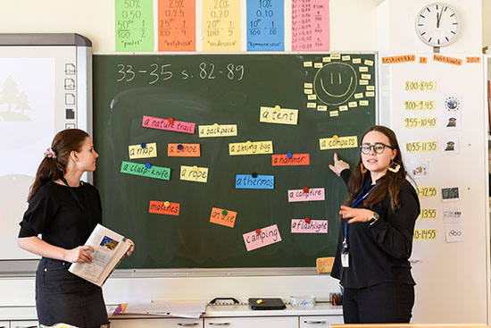 Kuvassa kaksi luokanopettaopiskelijaa opettavat liitutaulun edessä. 