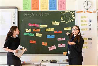 Kuvassa kaksi luokanopettaopiskelijaa opettavat liitutaulun edessä. 