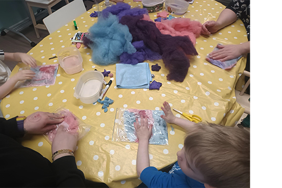 Pyöreä pöytä, jolla erilaisia askartelutarvikkeita ja jonka äärellä istuu lapsia ja aikuisia.