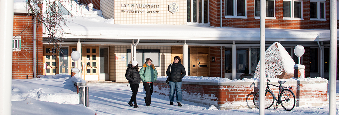 Kolme ihmistä kävelee Lapin yliopiston pääovien lähellä talvisena, aurinkoisena päivänä.