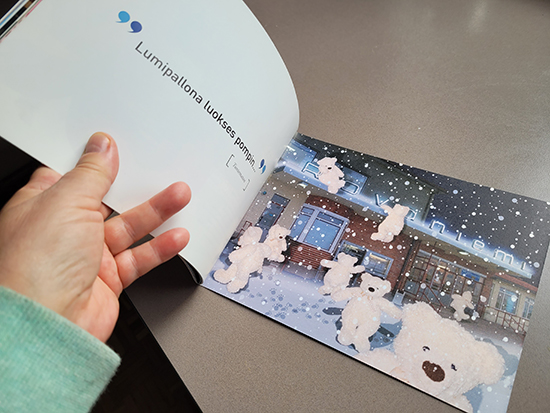 Käsi pitää avoinna kirjasta aukeamalta, jossa vasemmalla sivulla lukee Lumipallona luokses pompin ja oikealla sivulla on Jukka Kiviniemen kuvitus, jossa valkoisia nalleja seisoo rakennuksen pihalla ja kapuaa rakennuksen katolle.