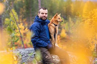Kuvassa Mikko Äijälä istuu ruskaisen maiseman keskellä pystykorva kainalossa.