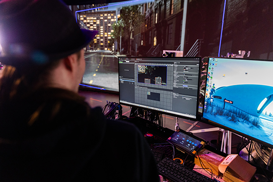 Virtuaalituotantostudion tietokonenäyttöjä ja lediseinä, joilla henkilö työskentelee selin kameraan. Kuva: Ville Rinne.