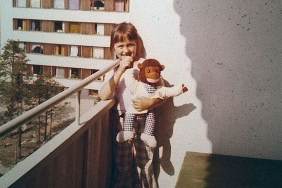 Parvekkeella seisoo tyttö, jolla on nalle kädessä. Taustalla kerrostalomiljöö.