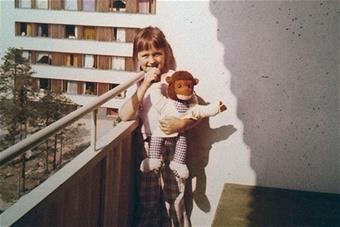 Parvekkeella seisoo tyttö, jolla on nalle kädessä. Taustalla kerrostalomiljöö.