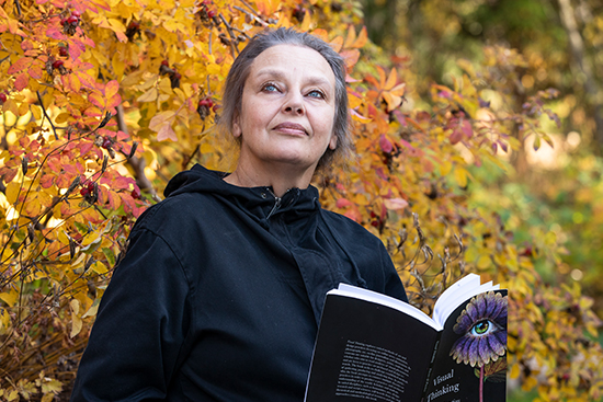 Leena Raappana-Luiro seisoo syksyisten pensaiden edustalla, katsoo yläviistoon ja pitää kädessään avonaista väitöskirjaansa.