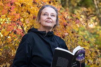 Leena Raappana-Luiro seisoo syksyisten pensaiden edustalla, katsoo yläviistoon ja pitää kädessään avonaista väitöskirjaansa.