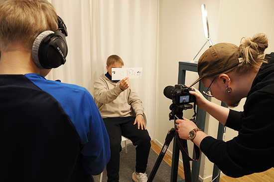 Lapsi istuu penkillä kuvausstudiossa ja pitää kasvojensa edessä korttia. Aikuinen katsoo häntä kameran kautta ja toinen lapsi seisoo kuvaajan vieressä kuulokkeet korvillaan.