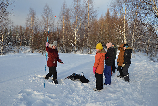 Tutkija näyttää mittatikun käyttöä koululaisille lumisessa maisemassa.