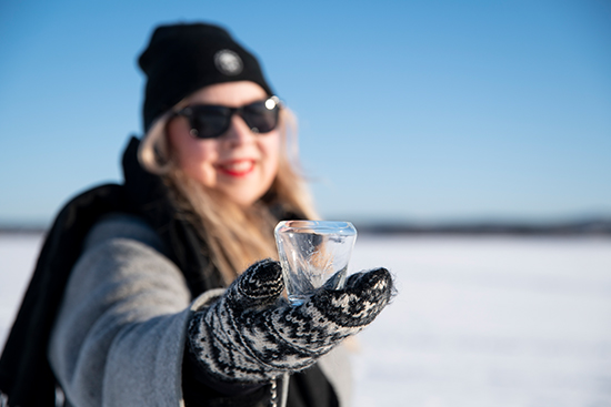 Milla Johansson pitää lapaseen puetussa, nostetussa kädessään suunnittelemaansa Kaltio-lasia. Kuva: Elli Alasaari.