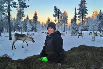 Korinna Korsström-Magga istuu kuivaheinien päällä talvisessa metsässä, taustallaan poroja. Kuva: Katariina Lehtonen.