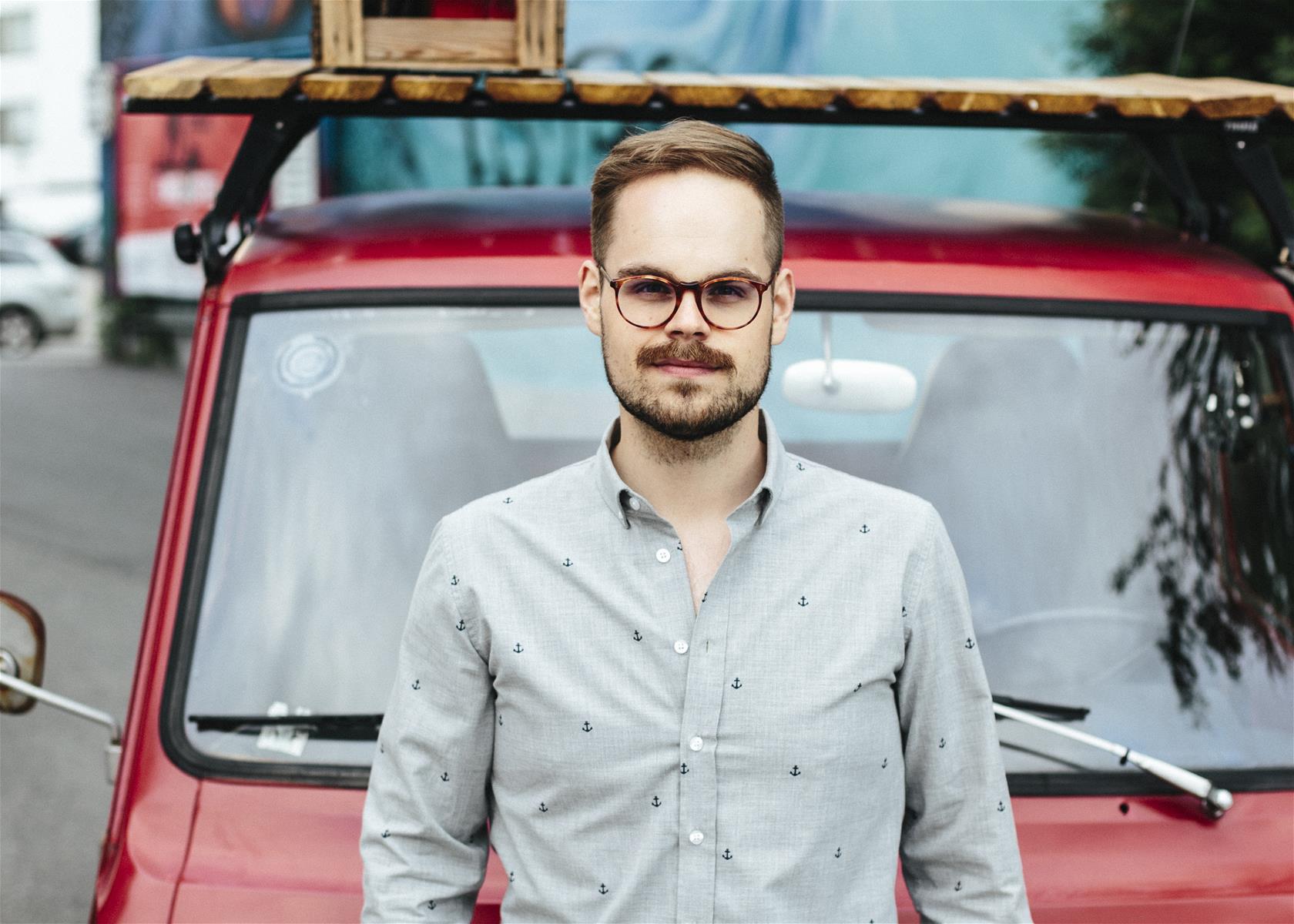 Silmälasipäinen mies punaisen auton edessä