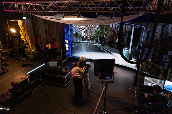 Yleiskuva Lapin yliopistolla sijaitsevasta virtuaalituotantostudiosta, jossa kaareva, iso lediseinä, kamera- ja valaistuslaitteistoa ja tietokoneita sekä henkilöitä työskentelemässä. Kuva: Ville Rinne.