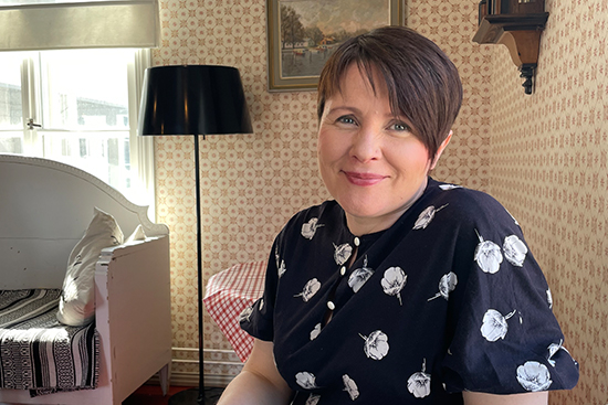 Maria Huhmarniemi istuu pöydän ääressä, taustalla kodin huonekaluja.