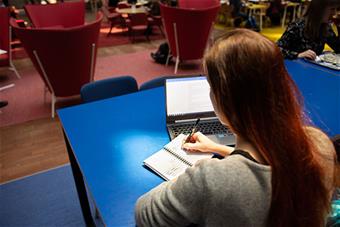 Kuvassa nuori nainen kirjoittaa muistiinpanoja tietokoneen äärellä. 