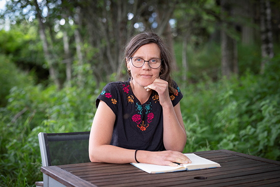 Kristiina Koskinen istuu pöydän ääressä. Hän nojaa leukaansa vasempaan käteensä, ja hänen oikea kätensä on pöydällä olevan avonaisen kirjan päällä. Taustalla on kesäisen vehreitä puita ja kasvillisuutta. Kuva: Harri Pälviranta.