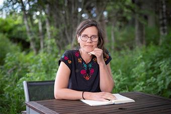 Kristiina Koskinen istuu pöydän ääressä. Hän nojaa leukaansa vasempaan käteensä, ja hänen oikea kätensä on pöydällä olevan avonaisen kirjan päällä. Taustalla on kesäisen vehreitä puita ja kasvillisuutta. Kuva: Harri Pälviranta.