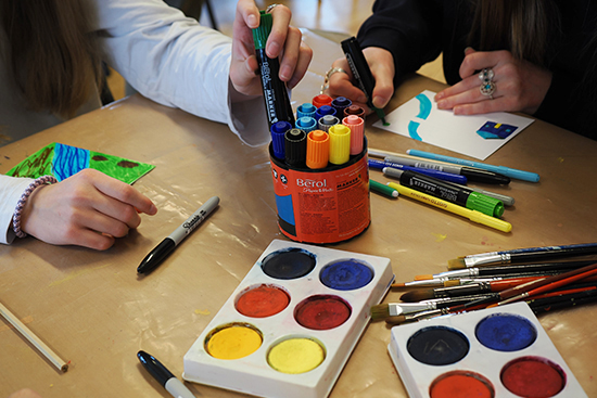 Tusseja ja vesiväripaletteja pöydällä, jonka äärellä istuu kaksi henkilöä. Toinen heistä piirtää korttiin ja toinen kurottaa kädellään tussia.