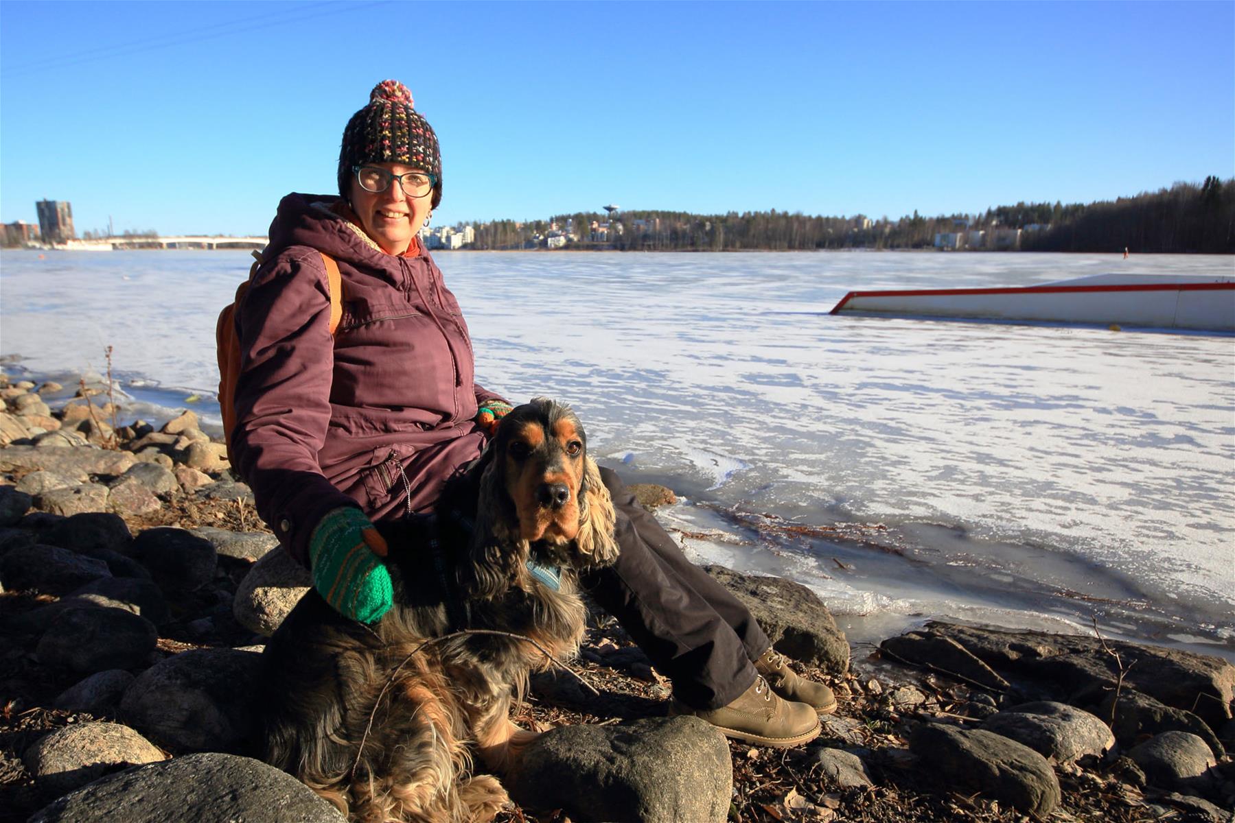 Kuvassa Riitta Penttinen istuu talvimaisemassa merenrannalla koiran kanssa. 