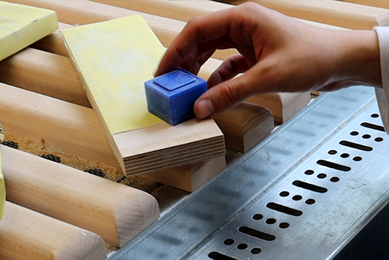 Käsi pitelee muovikuutiota puukappaleen yllä. Kuva: Siiri Paananen.