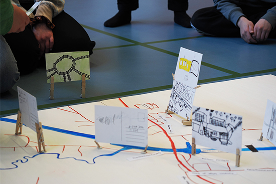 Lattialla olevan piirretyn kartan päällä on piirrettyjä kortteja, jotka on asetettu tietyille kartan alueille pyykkipojilla. Taustalla istuu lattialla ihmisiä kartan äärellä.