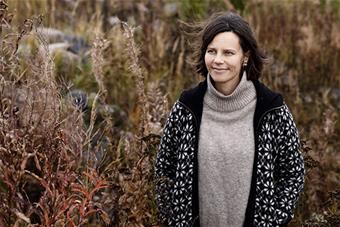 Kristiina Koskinen ulkona syksyisessä luonnossa, vierellään korkeita kasveja ja taustallaan kivikkoa. Kuva: Heidi Piiroinen.