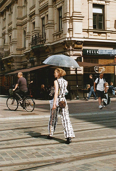 Kuva Sanni Kelokasken ja Venla Arkkon teos konseptista "Kelokaski & Arkko – Dance of the Cobblestones". Nainen kävelee raitiovaunukiskojen yli Tampereen kadulla, pidellen sateenvarjoa. Kuva: Venla Arkko.