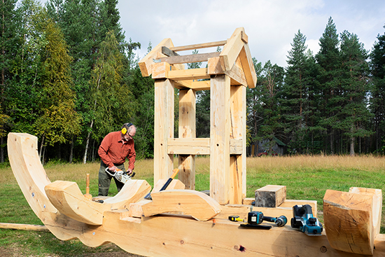 Timo Jokela veistää moottorisahalla hirsiveistosta, jossa veneen muotoa muistuttava vaakaosa ja sen päällä tornia muistuttava harjakattoinen rakennelma. Kuva: Aki Lintumäki.