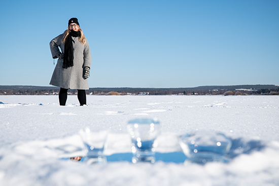 Milla Johansson seisoo matalassa lumihangessa Ounasjoen jäällä taustallaan vaarojen siluetti, etualalla kolme lasia jäällä. Kuva: Elli Alasaari.